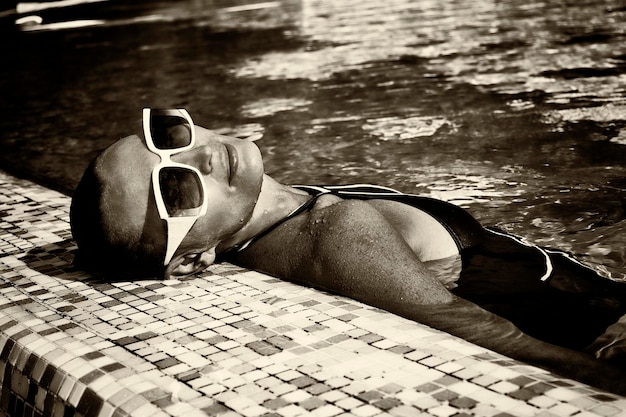 Foto vintage de uma mulher em um maiô preto e óculos de sol brancos nadando na piscina