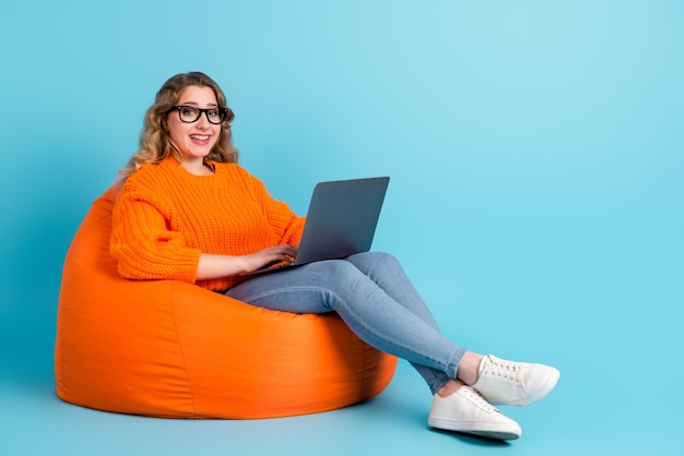 Foto vista de corpo inteiro de mulher digitando no laptop sentado na poltrona isolada em fundo colorido turquesa