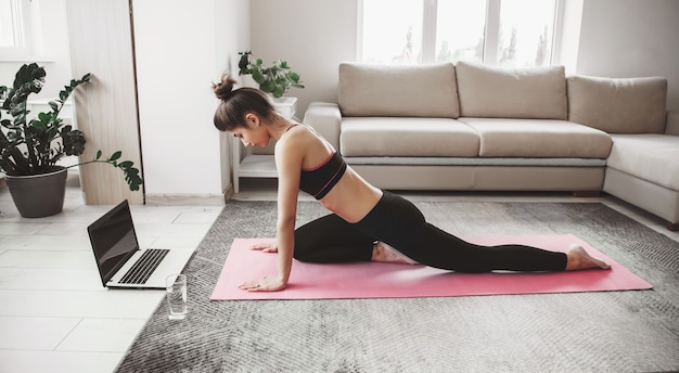 Foto de vista lateral de una dama caucásica practicando yoga en casa con una computadora portátil en el piso y un vaso de agua