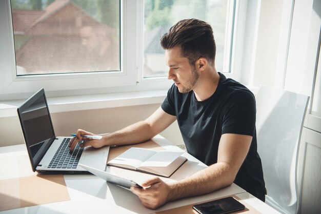 Foto de vista lateral de un hombre caucásico que trabaja en la computadora portátil desde casa con libro y tableta