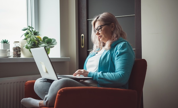 Foto de vista lateral de una mujer mayor caucásica escribiendo algo en la computadora portátil mientras usa gafas y ropa casual en un sillón