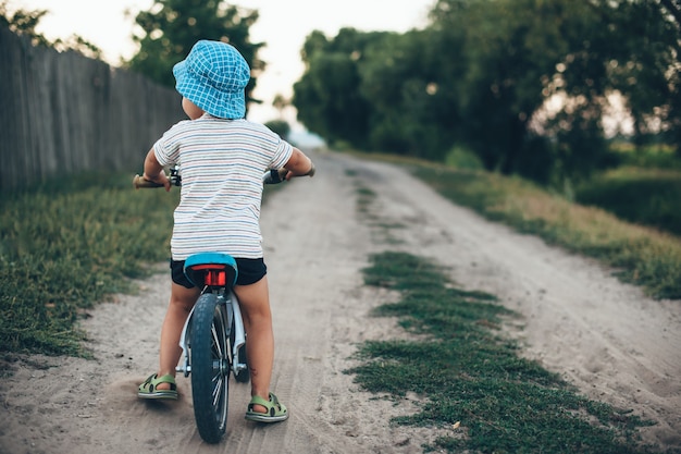 Foto de vista posterior de un niño en bicicleta con sombrero y caminando por una carretera rural