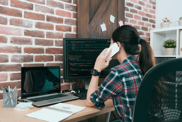 foto de vista posterior de una trabajadora de desarrollo de software de computadora bastante atractiva mirando el documento de código y usando llamadas de teléfono móvil con el cliente discutiendo el plan de diseño.