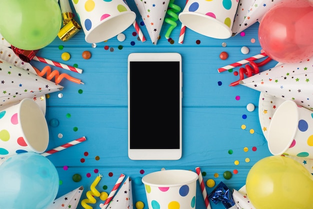 Foto de vista superior de accesorios de fiesta de composición de cumpleaños y teléfono inteligente con espacio de copia en el medio sobre fondo de mesa de madera azul aislado