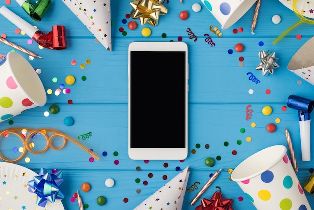 Foto de vista superior de accesorios de fiesta de composición de cumpleaños y teléfono inteligente con espacio de copia en el medio sobre fondo de mesa de madera azul aislado