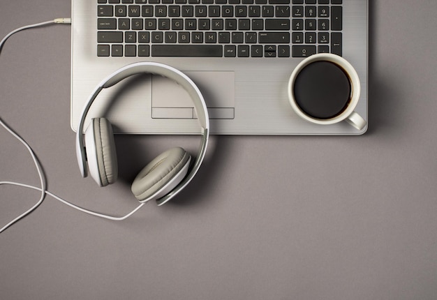 Foto de vista superior de auriculares blancos con cable y taza de café en el teclado de la computadora portátil sobre fondo gris aislado