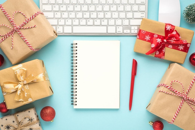 Foto de vista superior de bloc de notas, bolígrafo, papel artesanal, cajas de regalo, teclado, computadora, ratón, juguete de pino y bolas rojas de árbol de Navidad sobre fondo azul pastel aislado con espacio en blanco