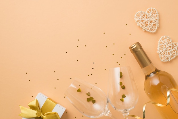 Foto de la vista superior de la caja de regalo blanca de la decoración del día de san valentín con lazo dorado botella de vino corazones de ratán copas de vino con confeti en forma de corazón y lentejuelas sobre fondo beige pastel aislado con espacio en blanco