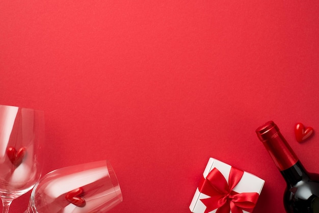 Foto de la vista superior de la caja de regalo blanca de las decoraciones del día de san valentín con corazones pequeños de lazo rojo en dos copas de vino y una botella de vino sobre fondo rojo aislado con espacio en blanco