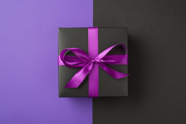 Foto de vista superior de caja de regalo negra con lazo de cinta morada sobre fondo violeta y negro bicolor aislado con espacio de copia