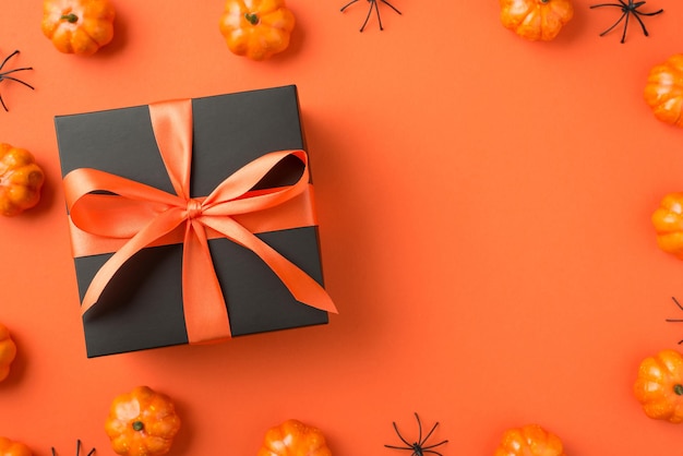 Foto de vista superior de caja de regalo negra con lazo de cinta naranja calabazas pequeñas y arañas sobre fondo naranja aislado con espacio en blanco