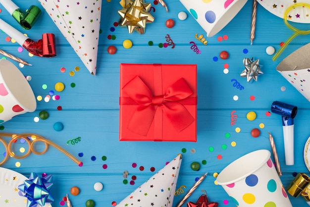 Foto de vista superior de caja de regalo roja con lazo de cinta en medio de la composición de cumpleaños y accesorios de fiesta sobre fondo de mesa de madera azul aislado