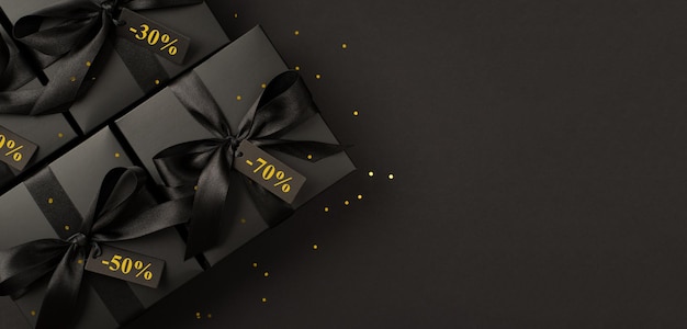 Foto de vista superior de cajas de regalo negras con lazo de cinta de raso negro y confeti dorado sobre fondo negro aislado con texto en etiquetas de precios