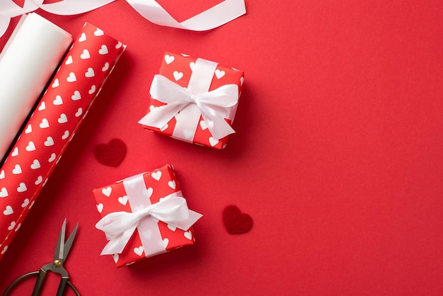 Foto de vista superior de cajas de regalo en papel de regalo rojo con patrón de corazones y lazos de cinta blanca corazones decorativos rollos de papel de regalo y tijeras sobre fondo rojo aislado con espacio vacío