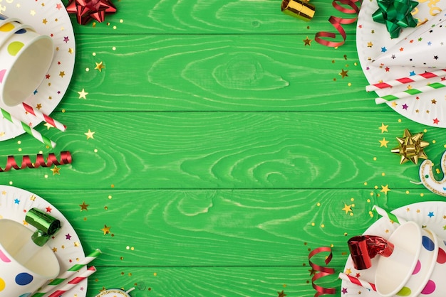 Foto de la vista superior de la composición de la fiesta de cumpleaños en las esquinas con cinta dorada verde roja estrellas sombrero lentejuelas tubos lunares vasos de papel y platos sobre fondo de mesa de madera verde aislado con copyspace