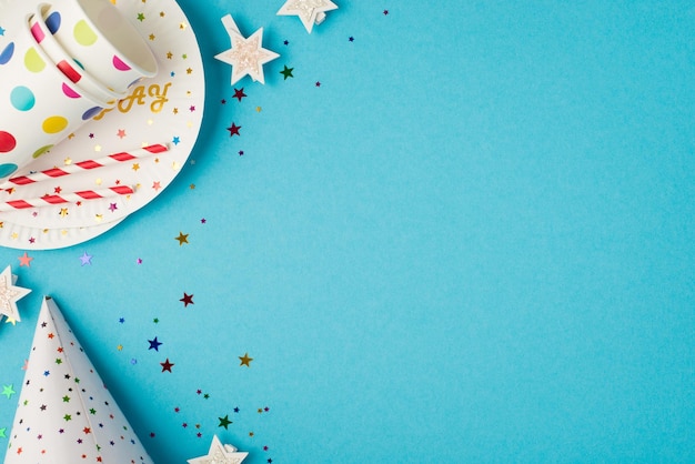 Foto de la vista superior de la composición de la mesa de cumpleaños con sombrero de lentejuelas, estrellas, vasos de papel y pajitas en un plato sobre fondo azul aislado con espacio de copia a la derecha