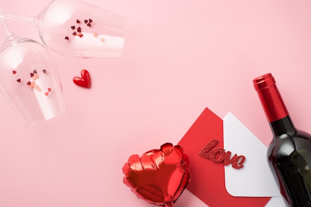 Foto de la vista superior de la decoración del día de san valentín dos copas de vino con confeti en forma de corazón globo sobre tarjeta roja inscripción amor y botella de vino sobre fondo rosa pastel aislado con espacio en blanco