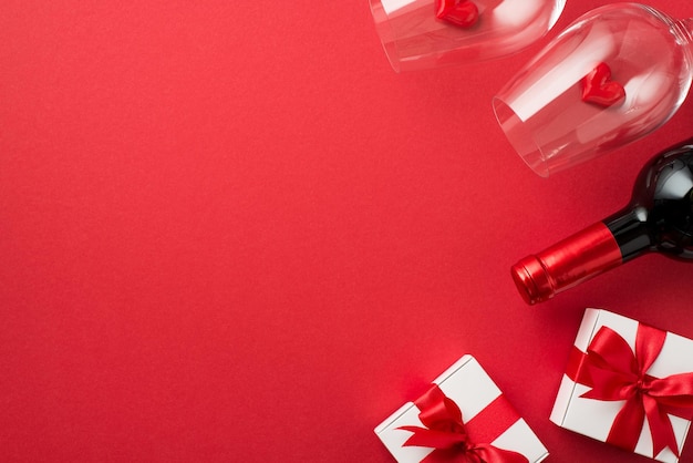 Foto de la vista superior de las decoraciones del día de san valentín cajas de regalo blancas con lazos de cinta roja pequeños corazones en dos copas de vino y una botella de vino sobre fondo rojo aislado con espacio vacío