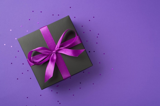 Foto de vista superior de una elegante caja de regalo negra con lazo de cinta púrpura y confeti sobre fondo violeta aislado con espacio de copia