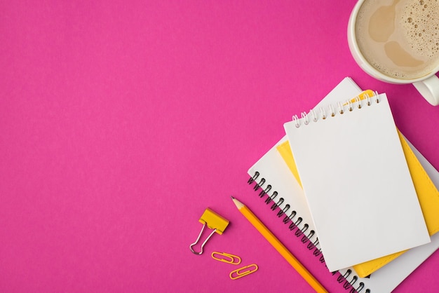 Foto de la vista superior del lugar de trabajo con cuadernos amarillos de la cancillería y una taza de café sobre fondo rosa aislado con espacio de copia