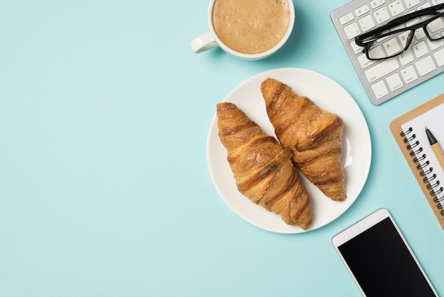 Foto de la vista superior del lugar de trabajo gafas de teclado portátil pluma teléfono móvil taza de café espumoso y plato con dos croissants sobre fondo azul pastel aislado con copyspace