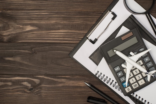 Foto de vista superior del lugar de trabajo con organizadores de carpetas negras de cancillería, calculadora de lupa, pluma y modelo de avión sobre fondo de mesa de madera aislado con espacio de copia