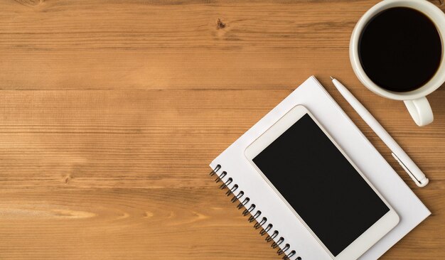 Foto de la vista superior del lugar de trabajo con una taza blanca de café y una pantalla de teléfono inteligente en el fondo de la mesa de madera aislada del cuaderno con espacio de copia