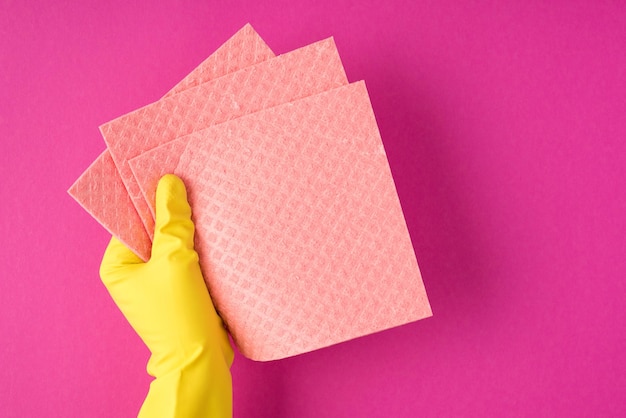 Foto de vista superior de la mano en guante amarillo sosteniendo trapos rosas sobre fondo rosa aislado con espacio de copia