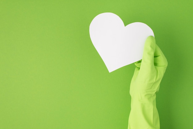 Foto de vista superior de la mano en guante de goma verde sosteniendo el corazón de papel blanco sobre fondo verde aislado con espacio en blanco