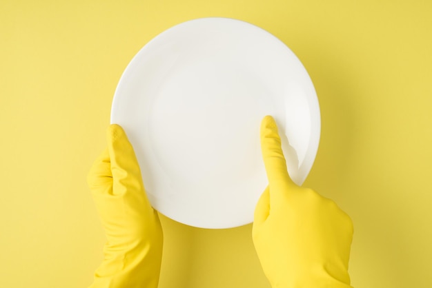 Foto de vista superior de manos en guantes de goma amarillos que indican con el dedo índice en un plato blanco claro sobre fondo amarillo aislado con espacio de copia