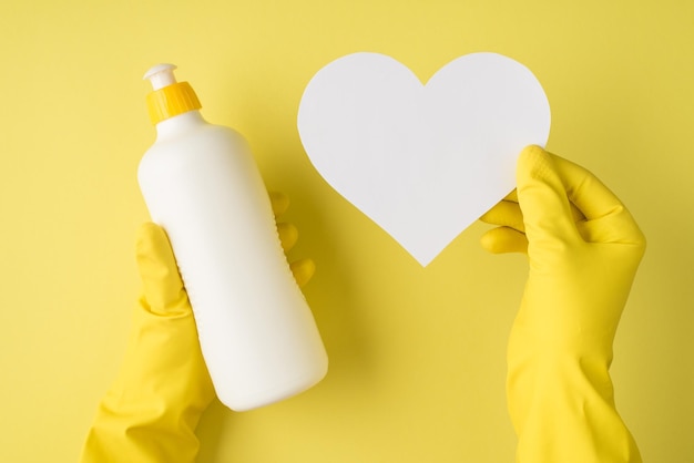 Foto de vista superior de manos en guantes de goma amarillos sosteniendo una botella de detergente blanco sin etiqueta y corazón de papel blanco sobre fondo amarillo aislado con espacio de copia