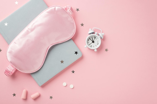 Foto de vista superior de máscara de dormir de seda rosa pequeñas píldoras de despertador blancas tapones para los oídos cuaderno azul y confeti en forma de estrella sobre fondo rosa pastel aislado con espacio vacío