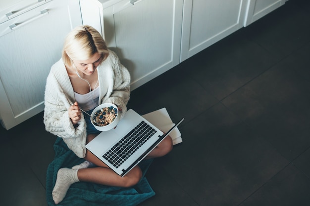 Foto de la vista superior de una mujer caucásica con cabello rubio y auriculares sentada en la cocina en el piso y usando una computadora portátil comiendo cereales