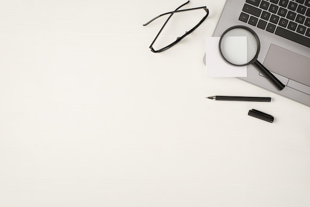 Foto de la vista superior del papel de nota de la etiqueta engomada de la lupa en el bolígrafo negro de la computadora portátil y gafas en el fondo de la mesa de madera blanca aislada con espacio vacío