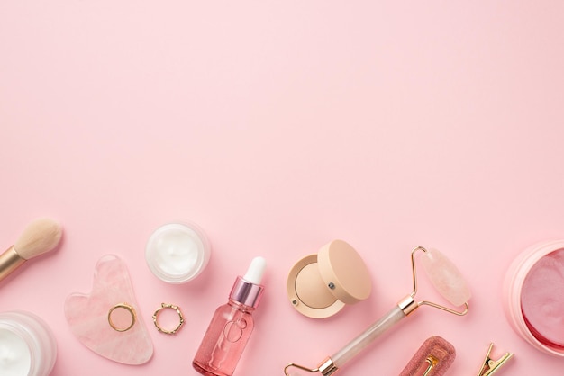 Foto de la vista superior del pincel de maquillaje rodillo de cuarzo rosa gua sha parches de ojos rosados botellas de vidrio horquillas de sombra de ojos y anillos de oro sobre fondo rosa pastel aislado con espacio de copia