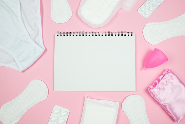Foto de vista superior del planificador abierto sobre toallas sanitarias, ropa de cama y pastillas sobre fondo rosa pastel aislado con espacio vacío