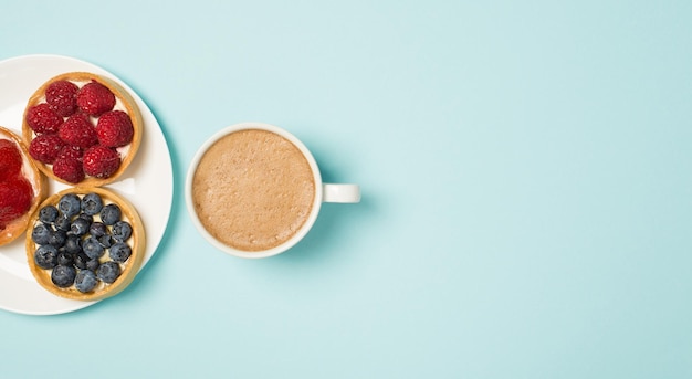 Foto de la vista superior del plato con tres pastelitos de bayas y una taza de café con leche espumoso sobre fondo azul claro aislado con espacio en blanco