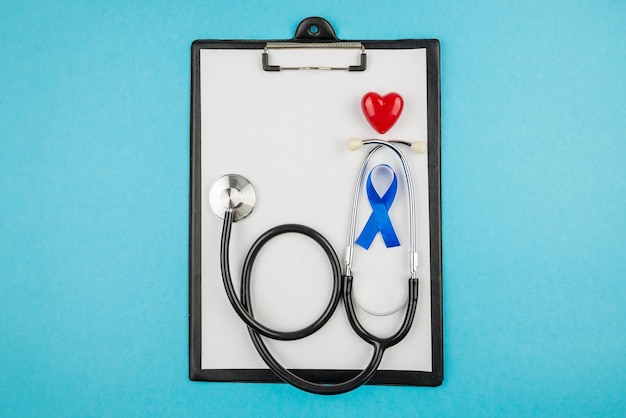 Foto de la vista superior del portapapeles del estetoscopio con una hoja de papel, cinta de seda azul y un pequeño símbolo de conciencia de cáncer de próstata de corazón rojo