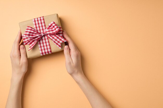 Foto de vista superior en primera persona de las decoraciones del día de san valentín manos femeninas tomando caja de regalo de papel artesanal con lazo de cinta a cuadros sobre fondo beige aislado con espacio vacío