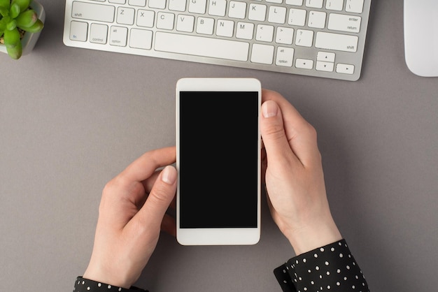 Foto de vista superior en primera persona de la maceta del ratón del teclado blanco y las manos de la mujer sosteniendo el teléfono inteligente en un fondo gris aislado con espacio de copia