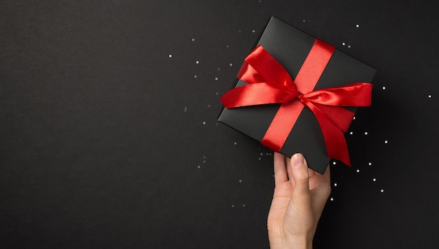 Foto de vista superior en primera persona de la mano femenina sosteniendo una caja de regalo negra con un arco de cinta roja vívida sobre lentejuelas brillantes en un fondo negro aislado con espacio vacío