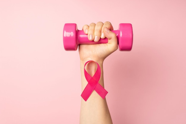 Foto de vista superior en primera persona de la mano de una mujer levantada sosteniendo una pesa rosa y una cinta rosa en la muñeca, símbolo de la conciencia del cáncer de mama en un fondo rosa pastel aislado