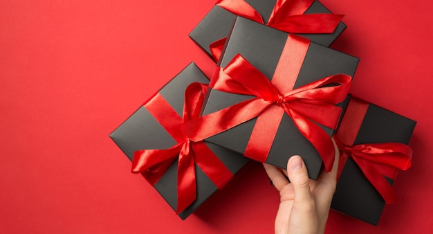 Foto de vista superior en primera persona de la mano sosteniendo una caja de regalo negra con un lazo de cinta roja sobre una pila de cajas de regalo en un fondo rojo aislado con espacio vacío