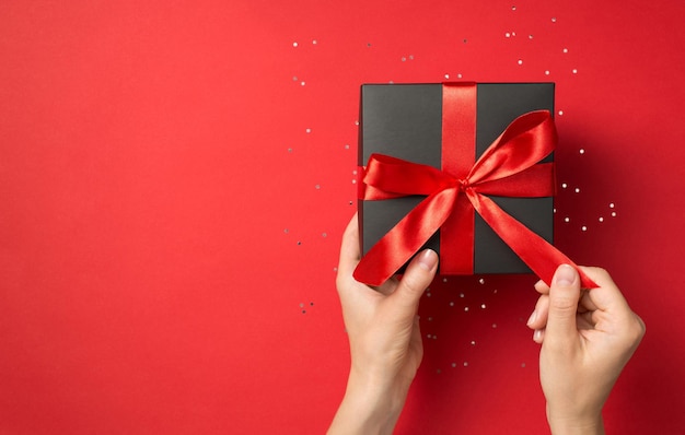 Foto de vista superior en primera persona de manos desempacando caja de regalo negra con lazo de cinta roja sobre fondo rojo aislado con espacio en blanco