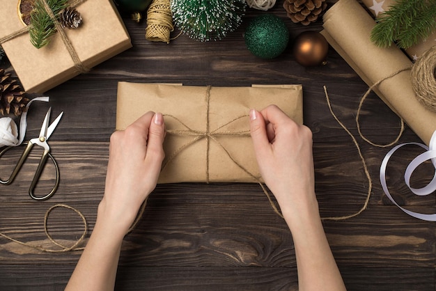 Foto de vista superior en primera persona de manos femeninas atando lazo de hilo en caja de regalo de paquete de papel artesanal bolas de oro verde ramitas de pino y herramientas artesanales en fondo aislado de escritorio de madera oscura