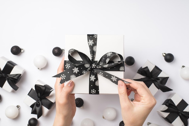 Foto de vista superior en primera persona de manos femeninas desempacando caja de regalo blanca con lazo de cinta negra sobre regalos bolas de árbol de Navidad blancas y negras sobre fondo blanco aislado con espacio de copia
