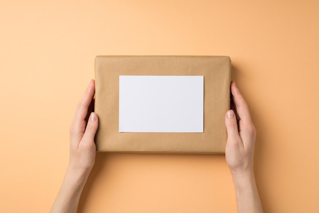 Foto de vista superior en primera persona de manos femeninas sosteniendo caja de regalo de papel artesanal con tarjeta de papel sobre fondo beige aislado con espacio de copia