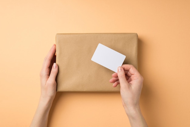 Foto de vista superior en primera persona de manos femeninas sosteniendo caja de regalo de papel kraft y tarjeta de plástico sobre fondo beige aislado con espacio en blanco