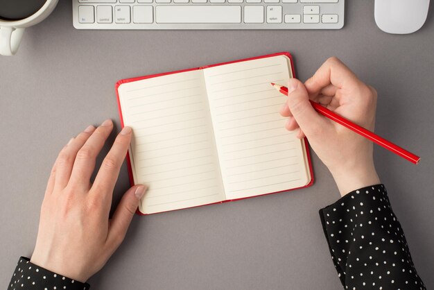 Foto de vista superior en primera persona de manos femeninas sosteniendo un lápiz sobre un organizador abierto rojo, un teclado blanco, un mouse y una taza de café en un fondo gris aislado con espacio en blanco