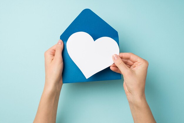 Foto de vista superior en primera persona de manos femeninas sosteniendo un sobre azul abierto con corazón de papel blanco sobre fondo azul pastel aislado con espacio en blanco